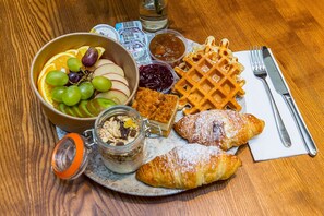 Bandeja de desayuno con waffles, croissants, un tazón de fruta y un frasco de mermelada.