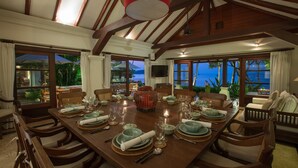 Un comedor con una mesa de madera lista para una comida, rodeado de sillas y grandes ventanas con vista al océano.