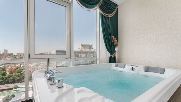 A modern bathroom with a large bathtub, a window with a city view, and a green curtain. - Rai Apartments (Odesa)