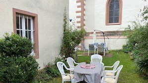 Um pátio com mesa e cadeiras, cercado por vegetação e um edifício com janela e porta.