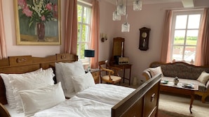 Quarto com cama de madeira, cômoda de madeira, sofá, escrivaninha com luminária, relógio e pintura de flores.