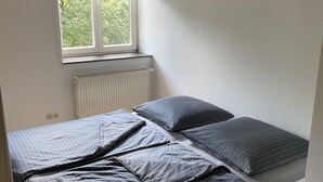A small, single-bed room with a window, a radiator, and a partial view of a green outdoor area.