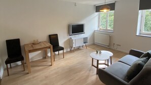 A spacious living room with a wooden floor, a grey sofa, a dining table with chairs, a TV on a white cabinet, and a window with a view of greenery.