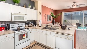 A modern kitchen with white cabinets, a microwave, stove, and a dishwasher. There is a window with a view of a pool and a ceiling fan. - Windsor Palms-ru556 (Kissimmee)