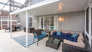 En uteplats med möbler i rotting, ett bord i glas och utsikt över en pool och ett hus i bakgrunden.