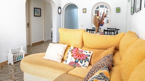 Un salon avec un canapé jaune, un fauteuil blanc et un coussin à motifs. Des tableaux encadrés ornent le mur, et un vase contenant des plantes séchées est posé sur une table.