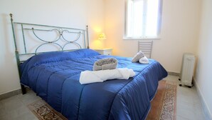 Een net opgemaakt bed met een blauwe deken, een opgerolde handdoek en een kussen op een metalen hoofdbord.