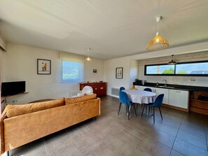 Un salon comprenant un canapé, une table à manger avec des chaises, un téléviseur, un coin cuisine avec une fenêtre et des ventilateurs de plafond.