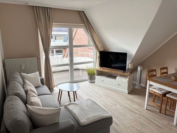 A modern living room with a sofa, TV, dining table, and a view of a brick building through a large window. - "Ruhekajüte" 2-room beachfront apartment in Büsum (Büsum)