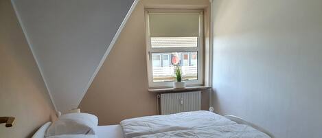 A single-bed room with a sloped ceiling, a window with a view of buildings, a radiator, and a bedside table with a lamp.