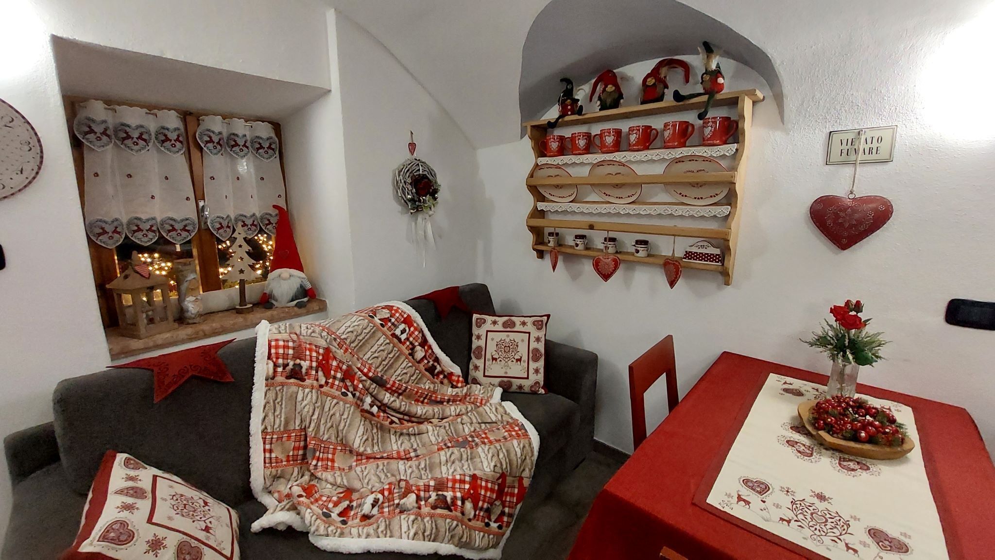 A cozy living room with a sofa, a table with a red tablecloth, and a shelf with decorative items.