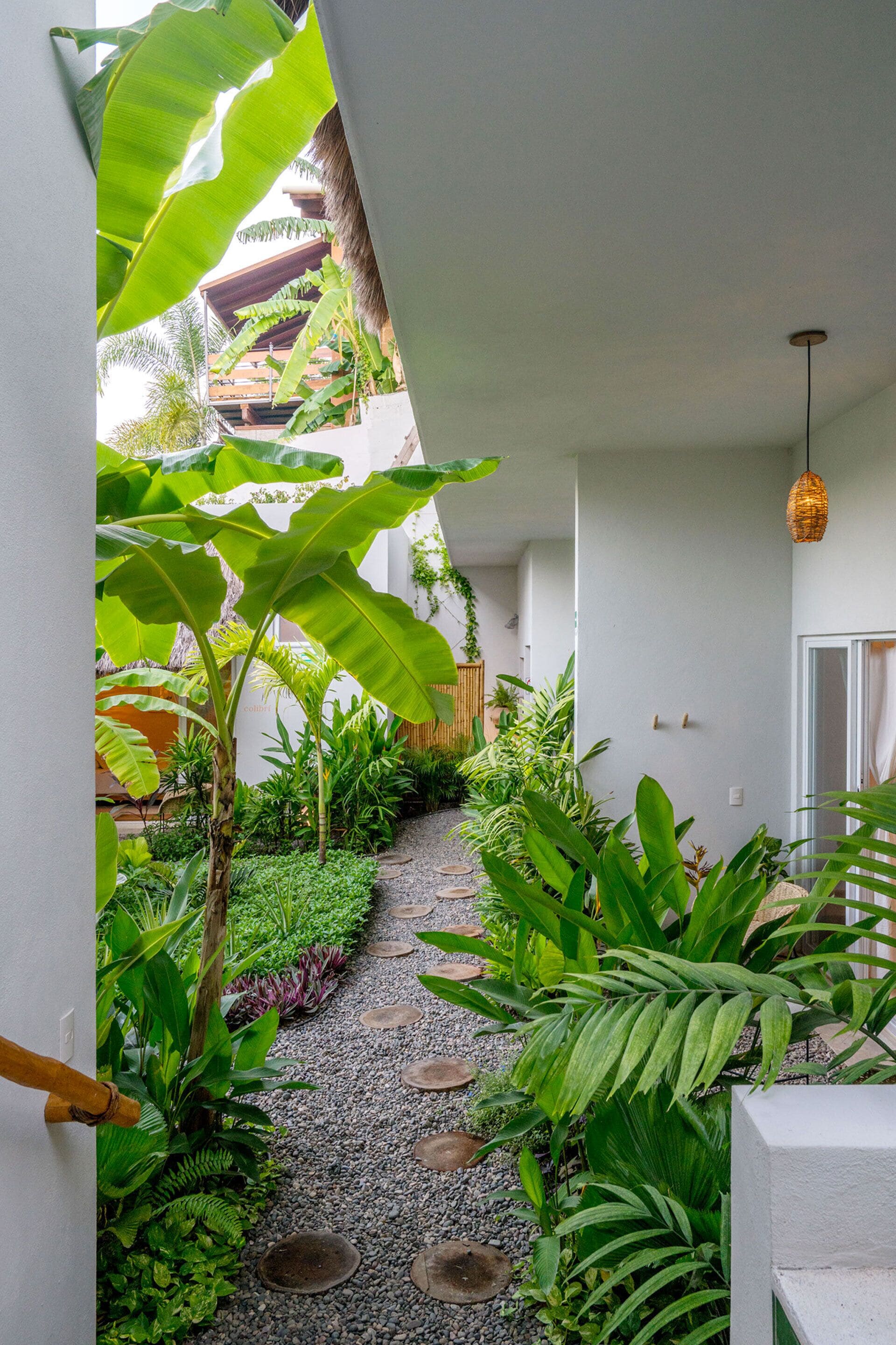 A narrow pathway flanked by lush tropical plants and a thatched-roof structure, leading to a white door.