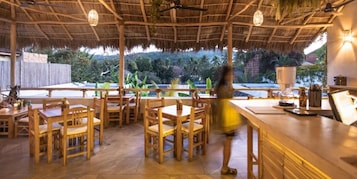 An outdoor dining area with wooden tables and chairs, a thatched roof, and a view of the surrounding landscape.