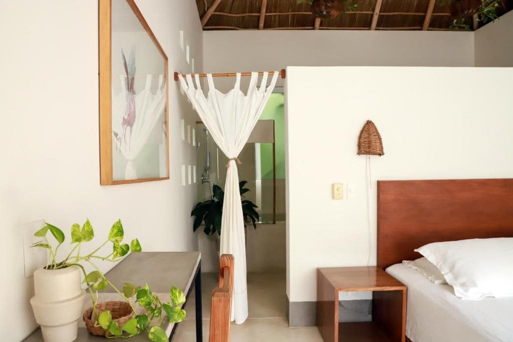 A room with a bed, bedside table, and a plant. There is a doorway leading to another room.