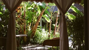 A room with a bed, white curtains, and a view of a lush garden with banana trees and a thatched roof structure.