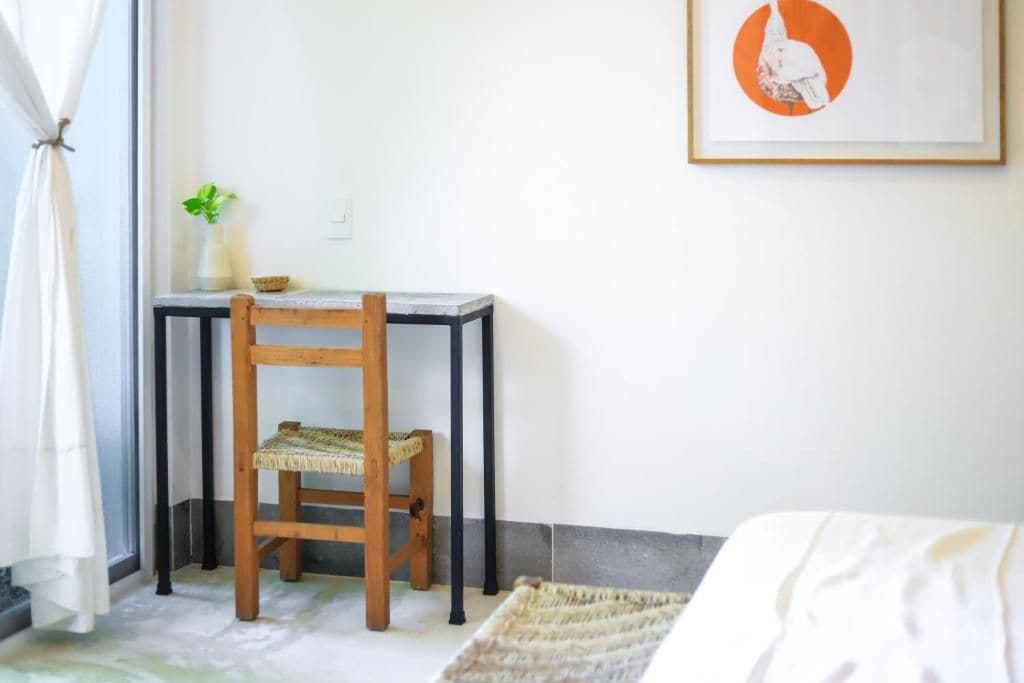 A room with a bed, a small table with a chair, a plant, and a framed picture on the wall.