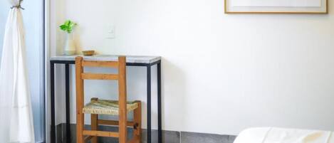 A room with a bed, a small table with a chair, a plant, and a framed picture on the wall.