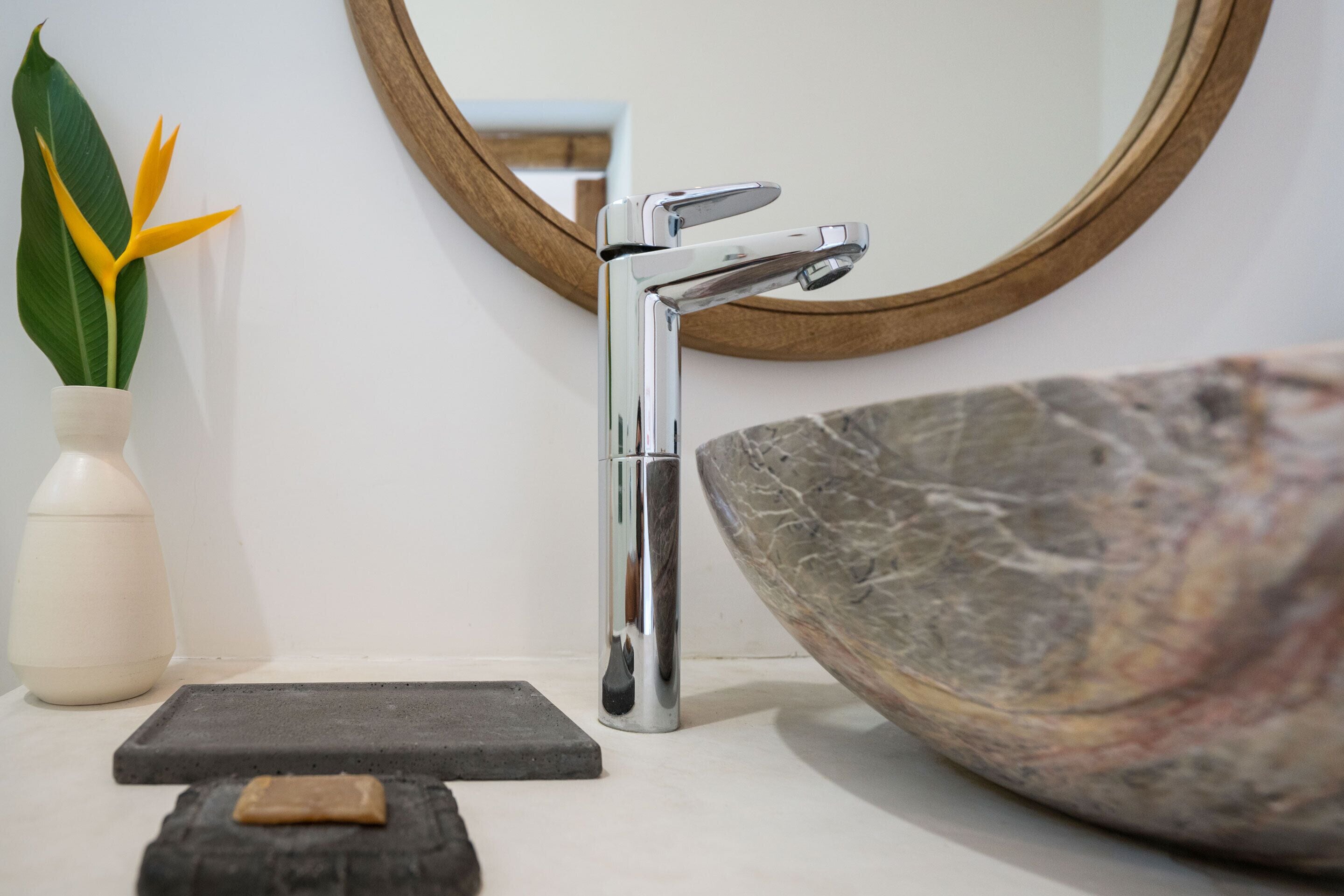 Un lavamanos de baño con un grifo moderno, un tazón decorativo grande, un jarrón pequeño con flores y un espejo con marco de madera.