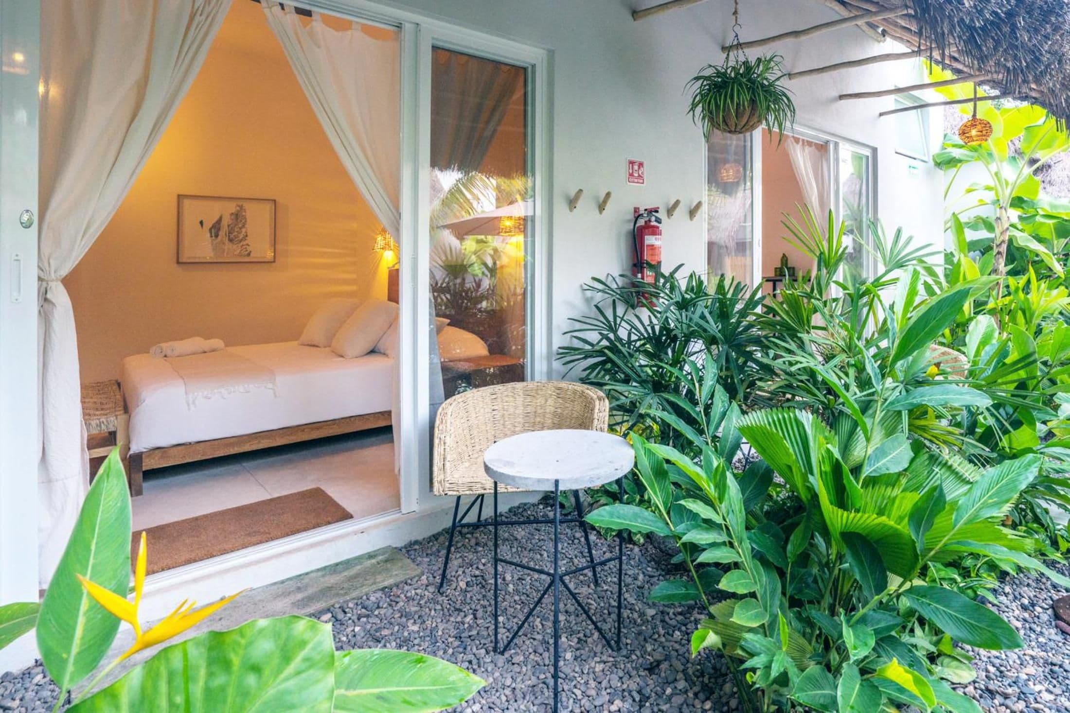 A room with a bed, a small table, and a chair, with a view of a garden outside.