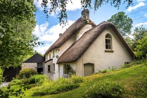 A traditional thatched-roof house with a stone foundation, surrounded by lush greenery and a clear sky.