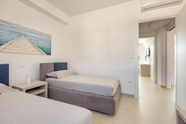A bedroom with two beds, a nightstand, a painting, and a hallway leading to another room.