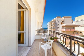 A balcony with a table and chairs, a potted plant, and a view of the sea and other buildings.
