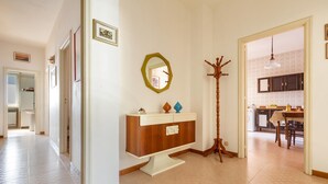 Un pasillo con un perchero de madera, una consola blanca con espejo y una zona de comedor visible a través de una puerta abierta.