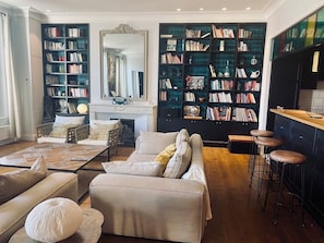 Ein Wohnzimmer mit Sofa, Couchtisch und Barhocker. Zwei große Bücherregale sind mit Büchern gefüllt. Im Hintergrund ist ein Kamin erkennbar.