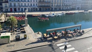A waterfront area with buildings, a dock with boats, and outdoor seating by the water.
