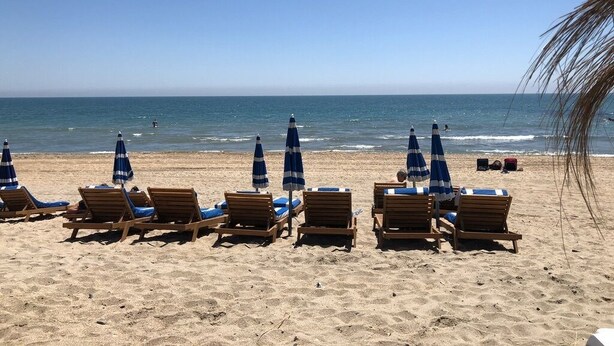 A beach with rows of wooden lounge chairs and blue umbrellas, facing the ocean.