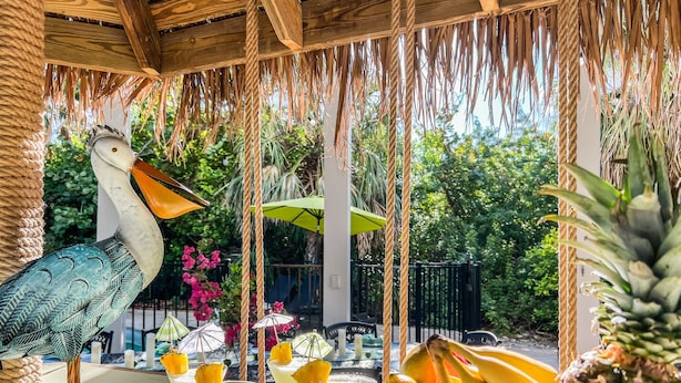 A tropical outdoor seating area with a wooden table, a decorative bird sculpture, and a basket of fruits. - Tropical Oasis - Pool w/ Waterfall & Swing Bar (Captiva)