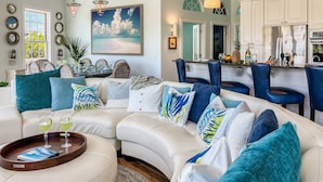 A modern living room with a white sectional sofa, blue and white patterned pillows, a dining area with wicker chairs, and a kitchen with white cabinets and blue bar stools. - Tropical Oasis - Pool w/ Waterfall & Swing Bar (Captiva)