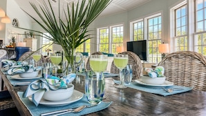 A dining area with a long table set for a meal, featuring a large potted plant, multiple glasses of beverages, and a television in the background. - Tropical Oasis - Pool w/ Waterfall & Swing Bar (Captiva)