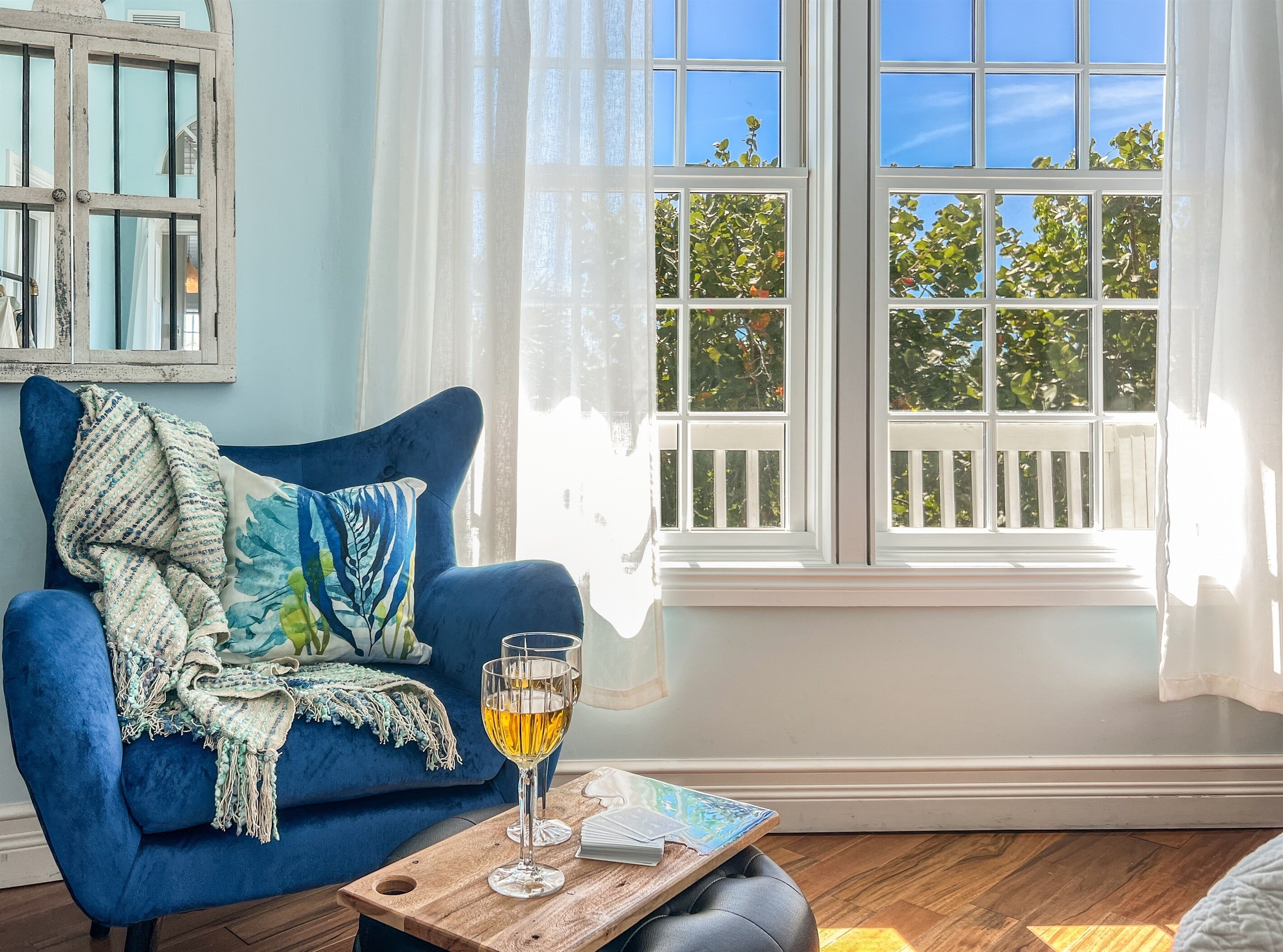 Una habitación acogedora con un sofá azul, una mesita de madera con una copa de vino, y vistas a árboles a través de un ventanal grande.