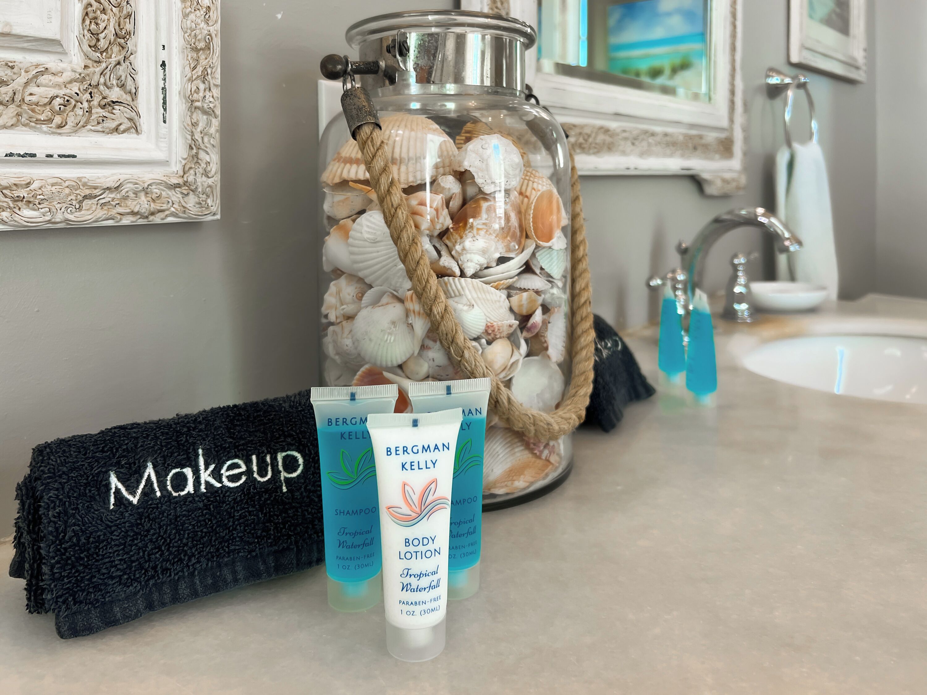 A bathroom with a glass jar filled with seashells, a black towel, and two tubes of Bergman Kelly body lotion and shampoo.
