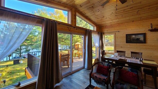 Une cabane en bois dotée de grandes fenêtres, d’un ventilateur de plafond, d’une table à manger et offrant une vue sur l’extérieur.