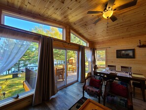 Eine Holzhütte mit großen Fenstern, einem Deckenventilator, einem Esstisch und Blick ins Freie.