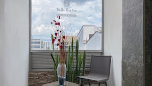 Un coin d’une chambre d’hôtel donnant sur la ville, un vase avec des fleurs rouges et un fauteuil gris.