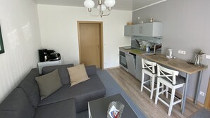 Un espacio habitable compacto con un sofá gris, una pequeña mesa de comedor con sillas, una kitchenette y una puerta de madera.
