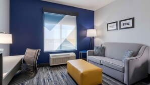 Ein modernes Hotelzimmer mit blauer Akzentwand, einem großen Fenster, einem grauen Sofa, einem gelben Ottomanen und einem Schreibtisch mit Stuhl.