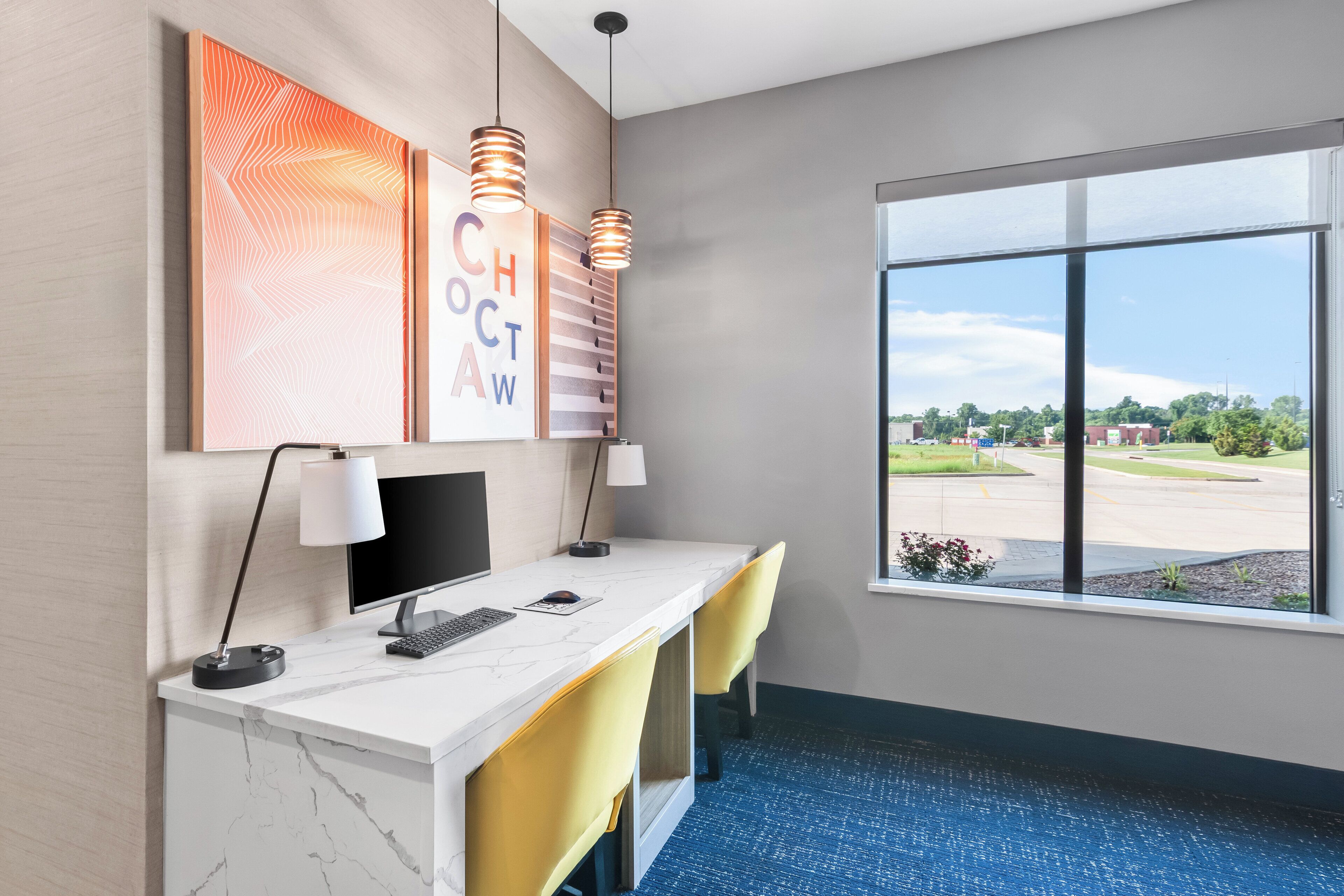 A hotel room with a desk, two chairs, a computer, and a large window offering a view of the parking lot and greenery.