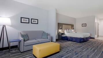 A hotel room with a grey sofa, a yellow ottoman, a bed with a blue bed skirt, a nightstand, a lamp, and two framed pictures on the wall.