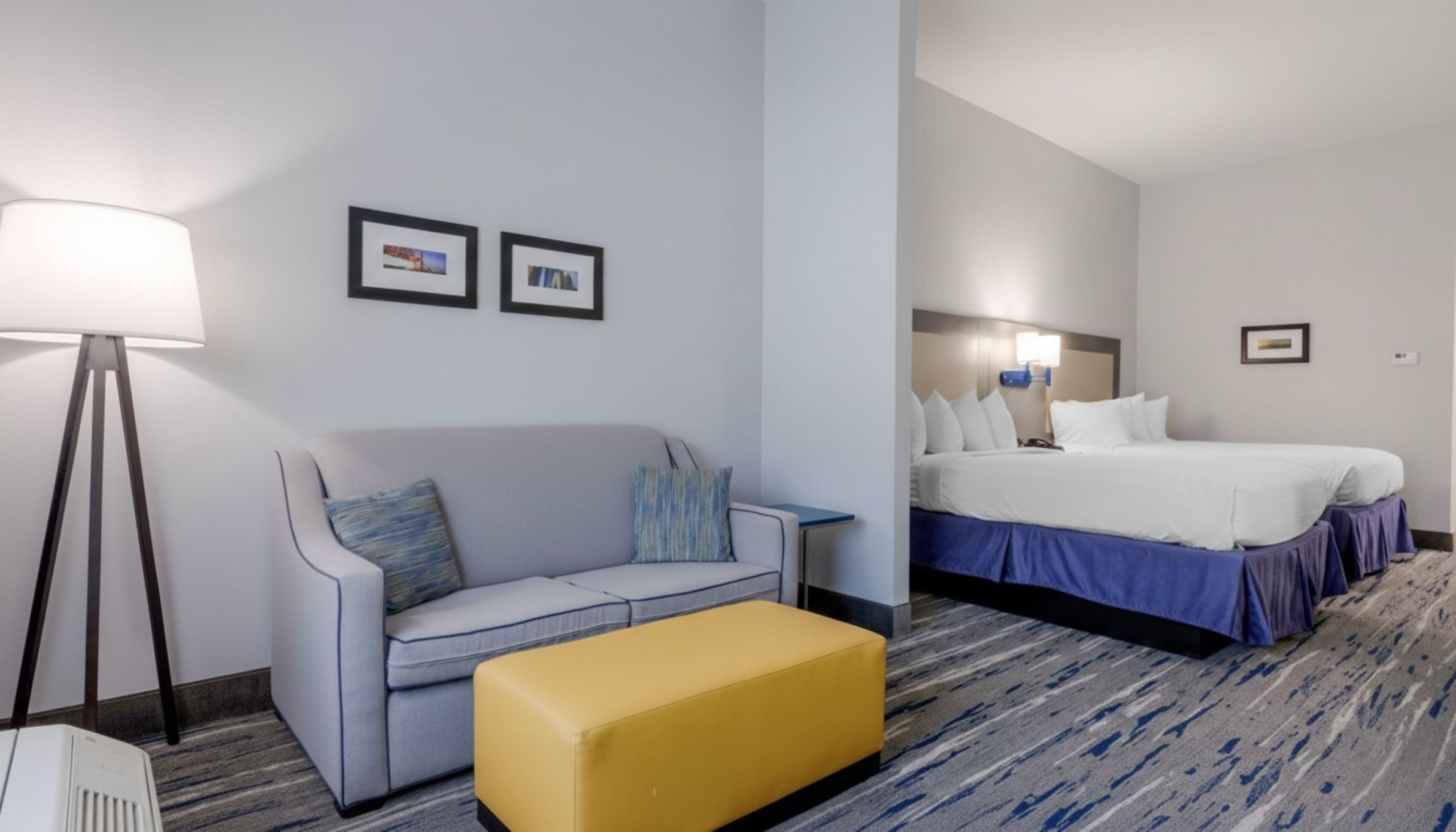 A hotel room with a sofa, a yellow ottoman, a bed with multiple pillows, a lamp, and two framed pictures on the wall.