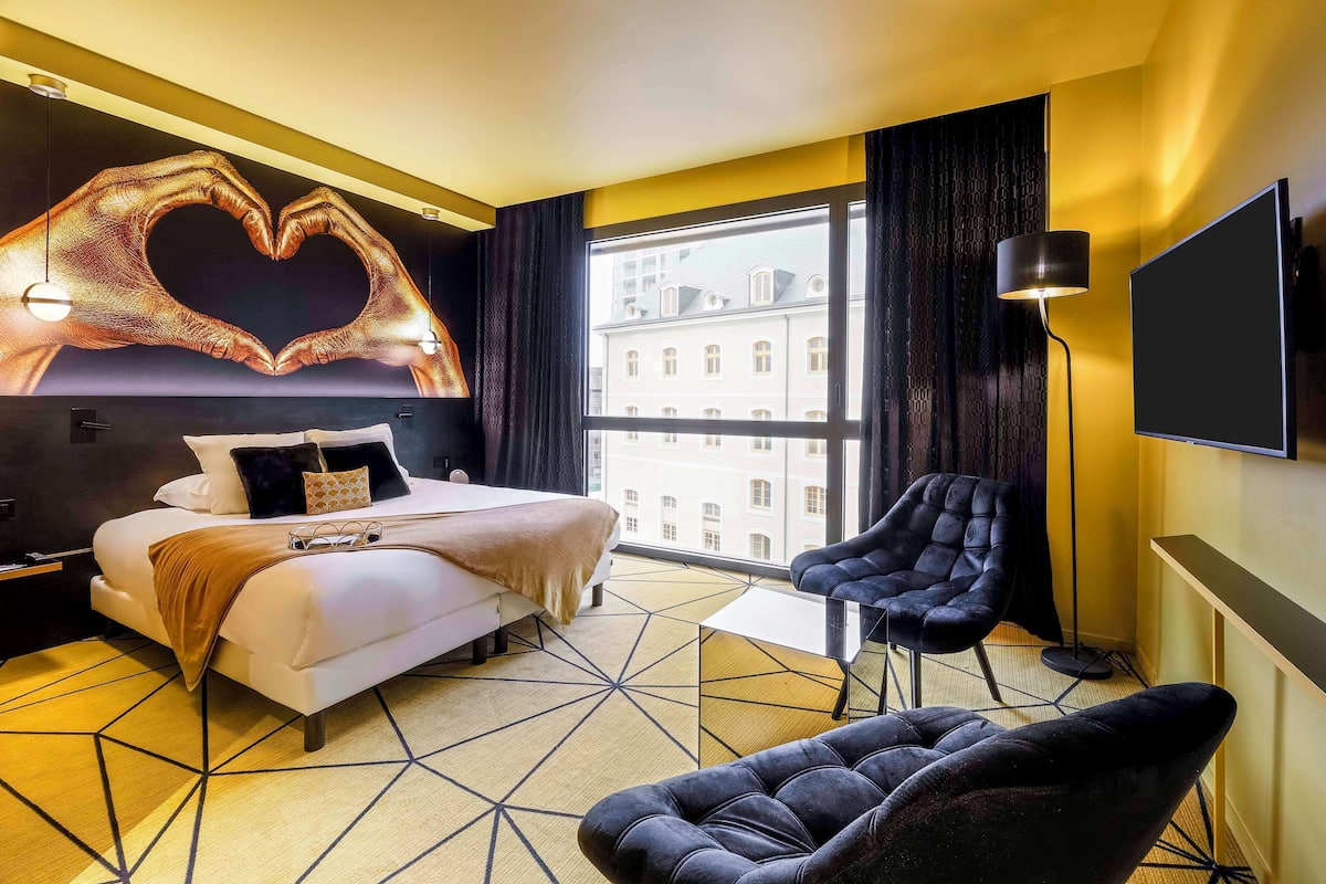 A modern hotel room with a large bed, a heart-shaped wall decoration, a black armchair, and a large window offering a city view.