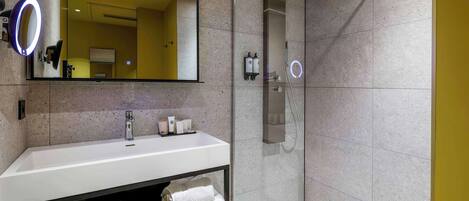 A modern bathroom with a white sink, a large mirror with a ring light, a glass-enclosed shower, and a vanity with towels.