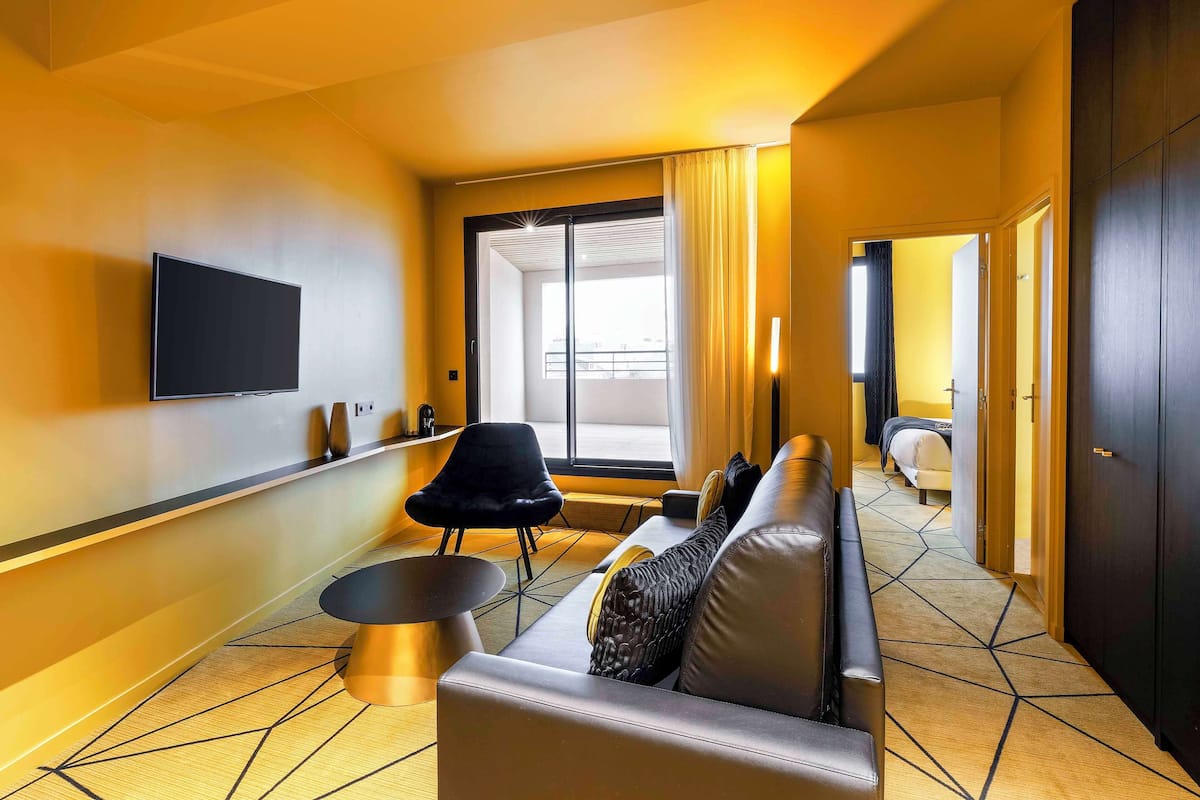 A modern hotel room with a sofa, a black chair, a flat-screen TV, and a large window with curtains.