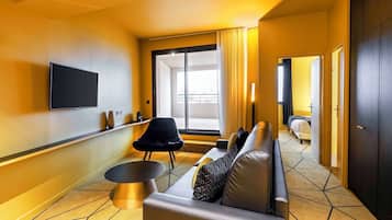 Ett modernt hotellrum med en soffa, en svart stol, en platt-TV och ett stort fönster med gardiner.