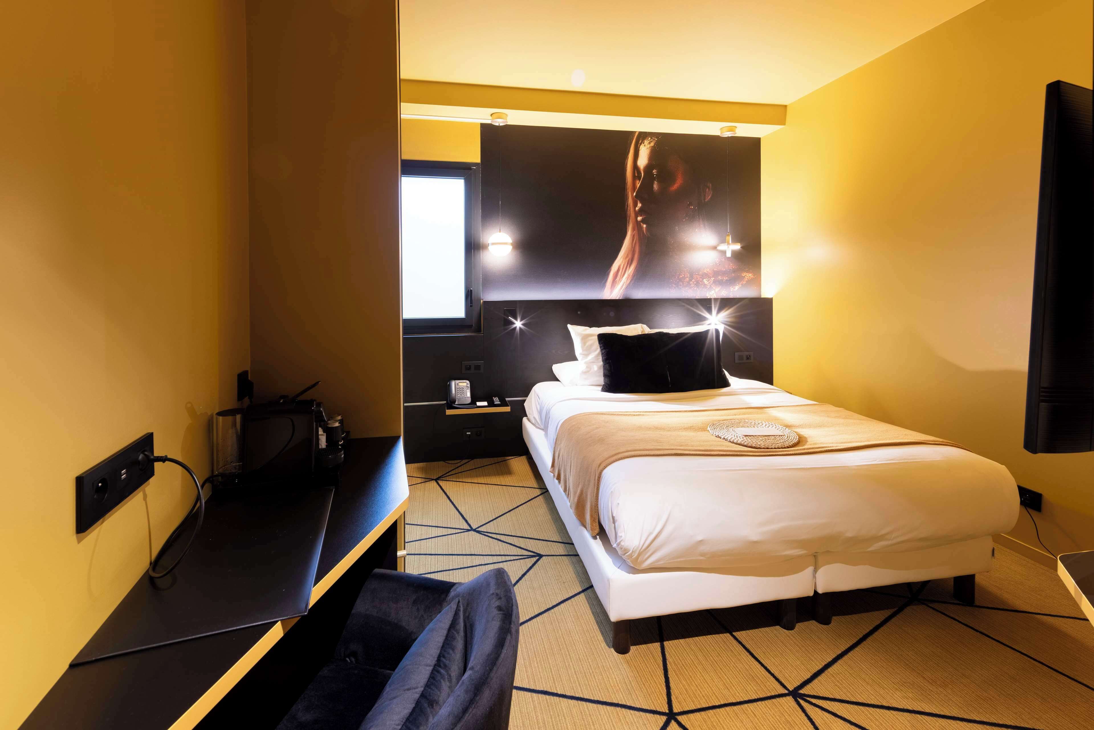 A hotel room with a bed, bedside table, lamp, and a large poster on the wall.
