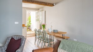A dining area with a wooden table and chairs, a view of the garden through an open door, and a cozy seating area with a sofa and armchair.