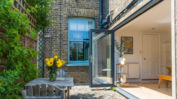A wooden bench with a vase of yellow flowers, a brick wall, and a glass door leading to a room with a white door and a painting. - Fig Tree House, Deal, Kent (Deal)